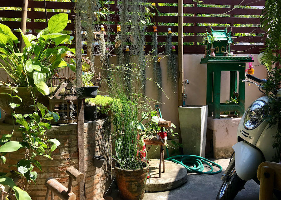 A sunny garden in Santitham with a Thai spirit house, plants, sculptures, and a motorbike.
