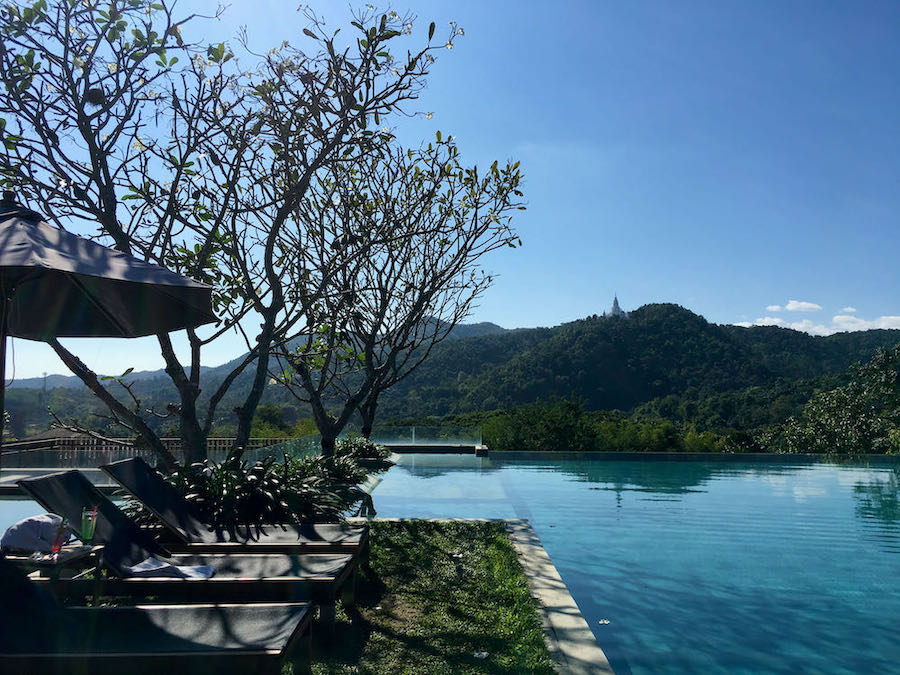 Infinity pool and lounge chairs at hillside resort with Chiang Mai hills and temple in background.