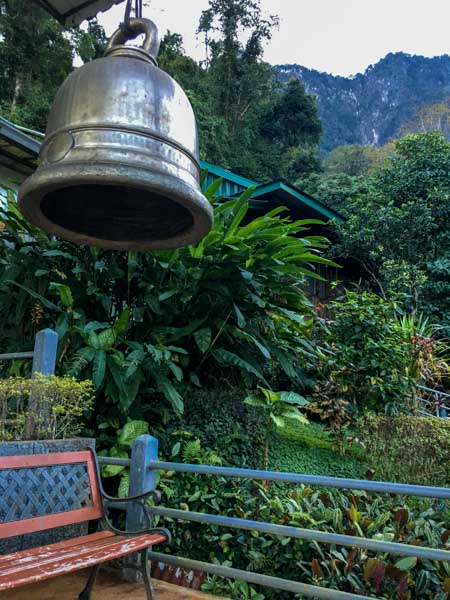 Large bell and bench surrounded by plants and mountains at Wat Tham Pha Plong.