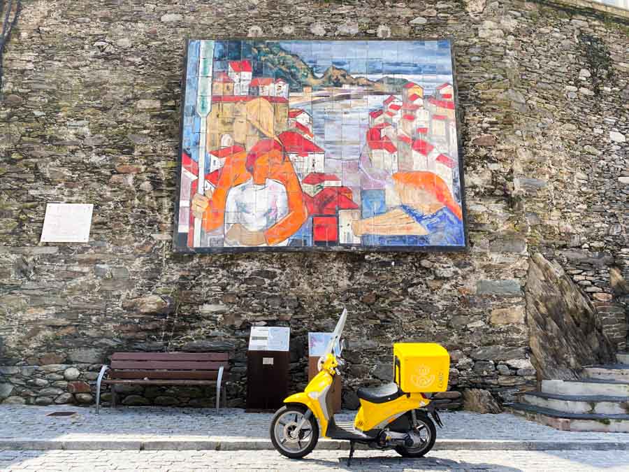 Painting of Cudillero rooftops and harbor, with a yellow Correos scooter parked in front.