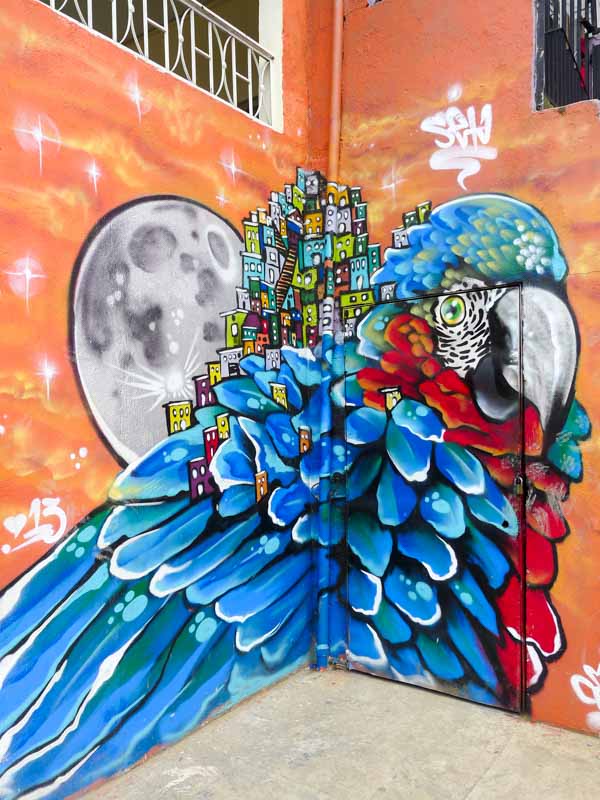 Colorful street art in Comuna 13 depicting a parrot, the moon, and Medellín's typical buildings.