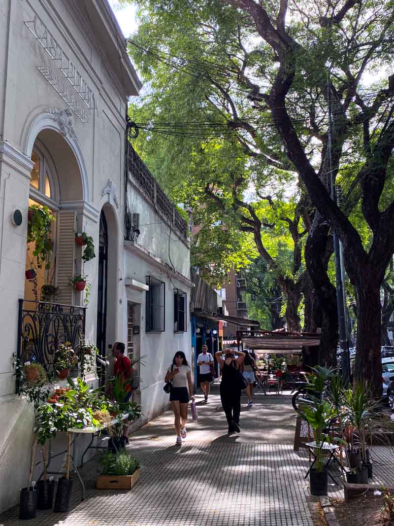 People walk past a flower shop on a leafy street with tall green trees in the Palermo barrio of Buenos Aires.