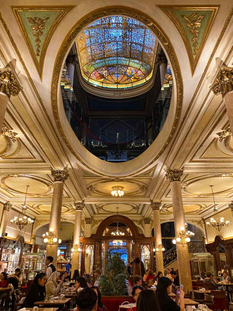 The gorgeous restored interior of Confitería La Ideal. People are enjoying cakes and coffee under the colorful stained glass ceiling.
