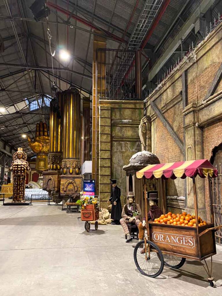 An orange cart and flower cart from the set of La Bohème with large gold sculptures from the set of Don Carlo in the background at Colón Fábrica.