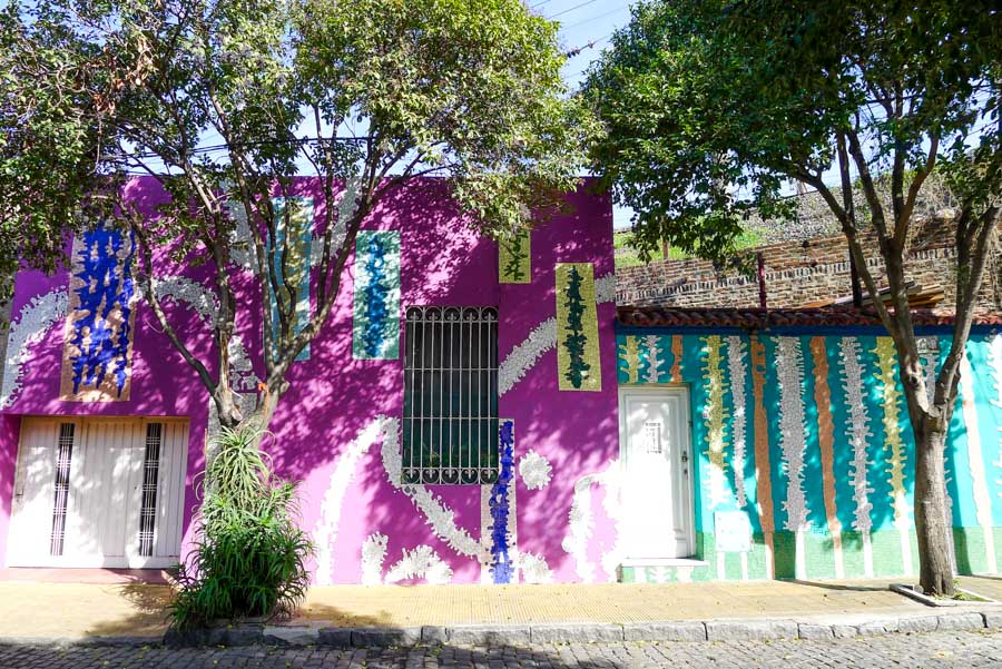 Trees cast pretty shadows on purple and teal facades decorated with multicolored mosaic tiles on Pasaje Lanín.