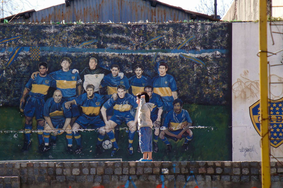 A young boy cheers next to a mural of the Boca Juniors soccer team.