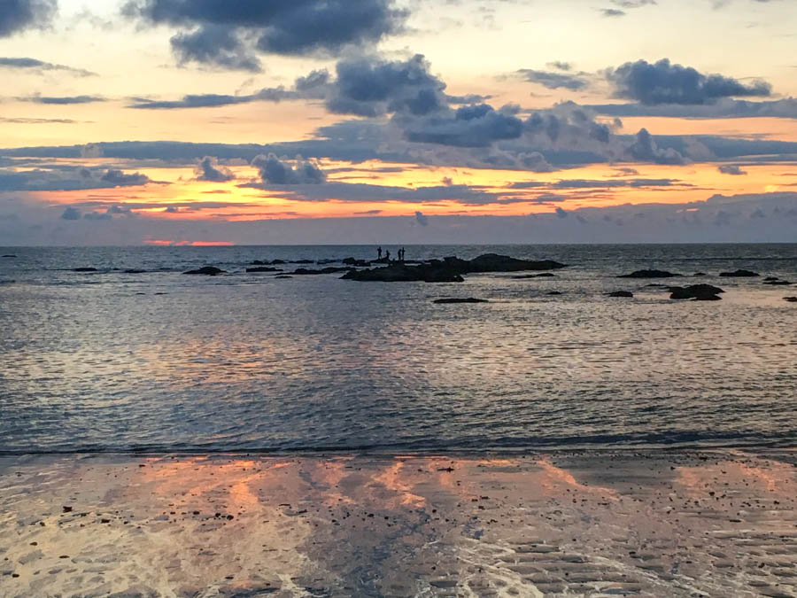 Four fishermen standing on distant rocks as the sun sets over the ocean in Khao Lak, Thailand.