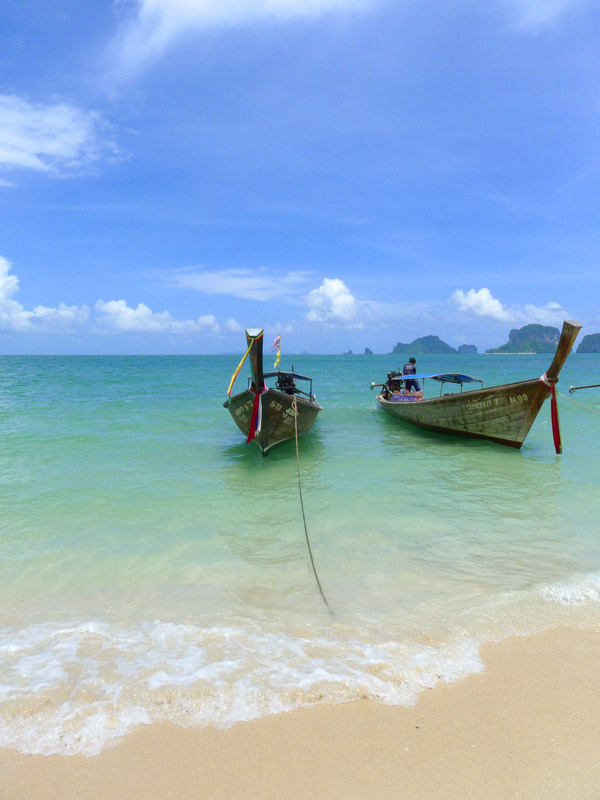 Distant limestone cliffs behind two longtail boats in clear turquoise water on Phra Nang Beach - one of the prettiest places on Thailand's southern Andaman coast.