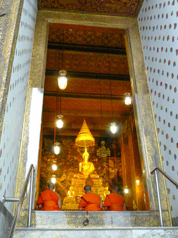 Three monks in orange robes sitting in front of a golden seated Buddha statue in the ordination hall of Wat Pho in Bangkok, a must-see on this 3-week Thailand itinerary.