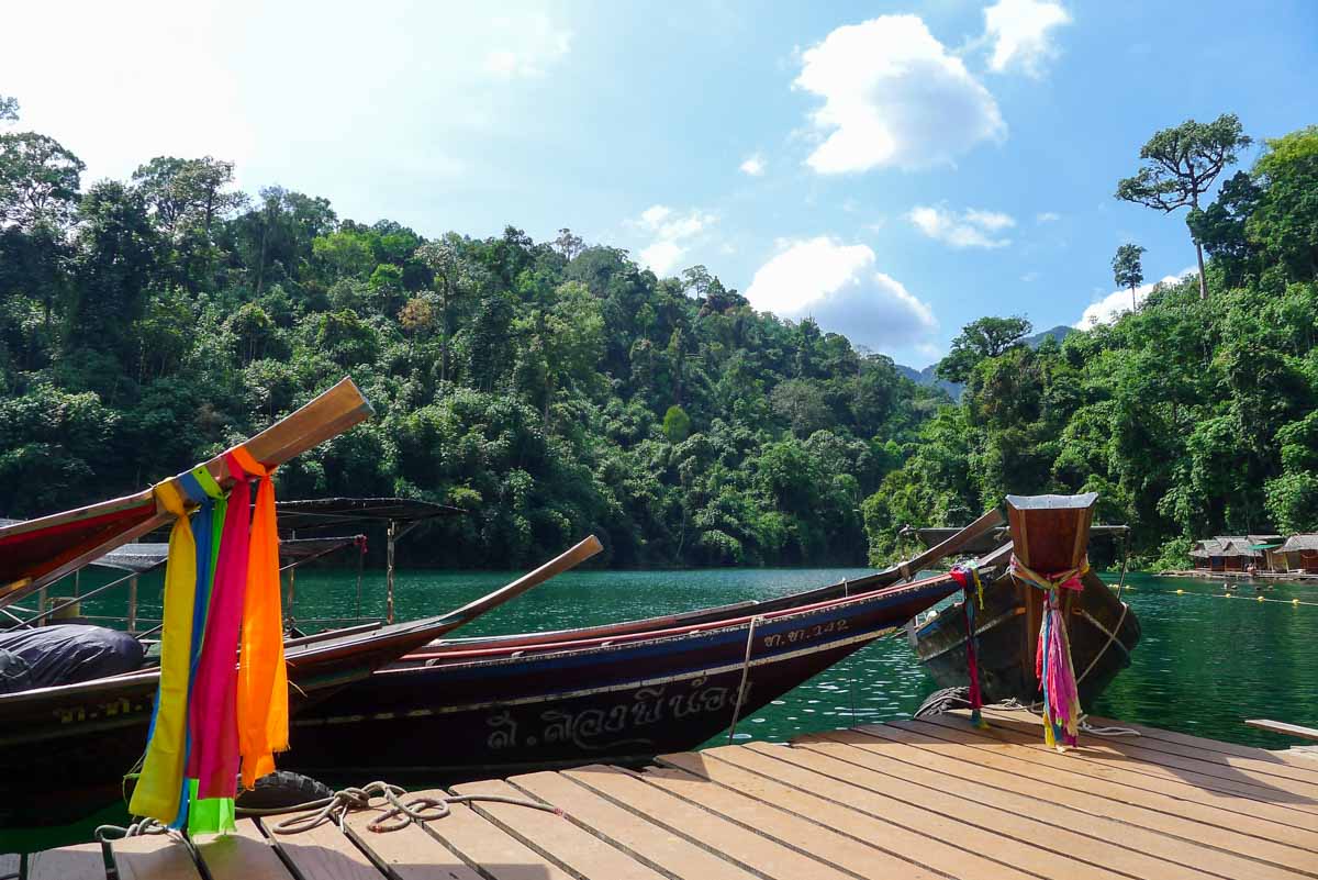 Classic Thai longtail boats decorated with brightly-colored fabric and moored to a wooden dock with a row of basic floating bungalows in the background.