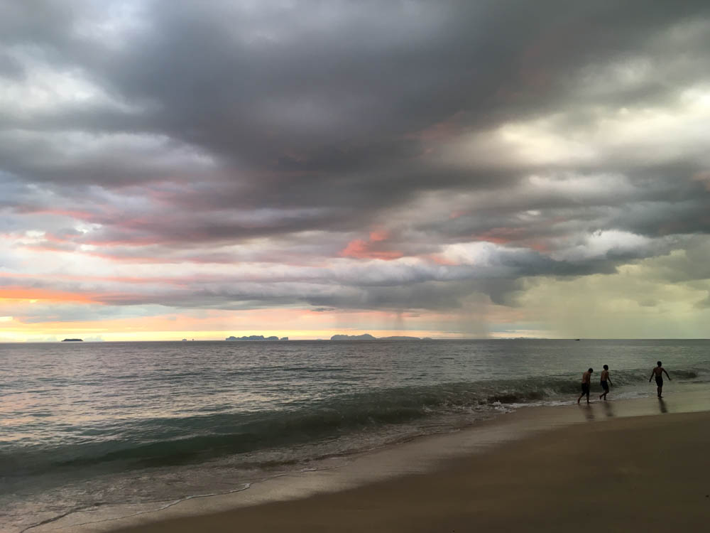 Three people walking in the waves under stormy skies on Koh Lanta. The sunset is just visible through the clouds but there are also rainstorms in the distance.