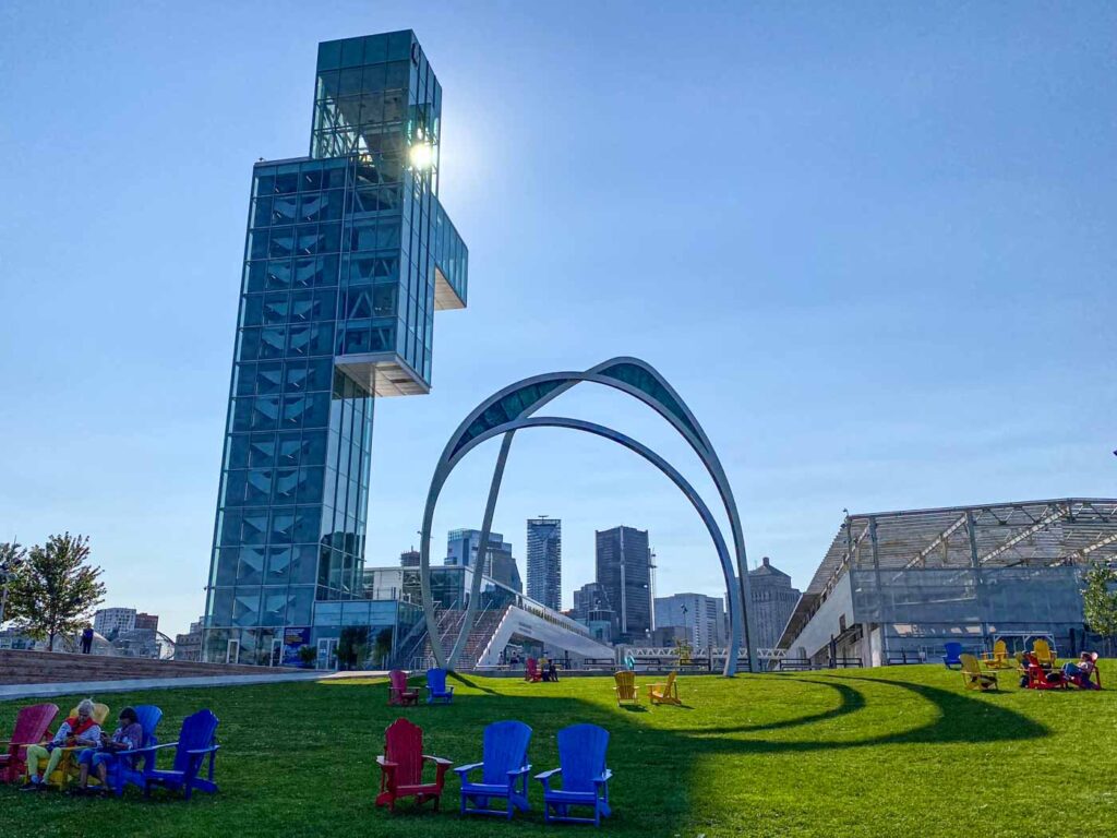 Blue, red, and yellow Adirondack chairs and a large sculpture in the grass next to the glass Port of Montreal Tower.