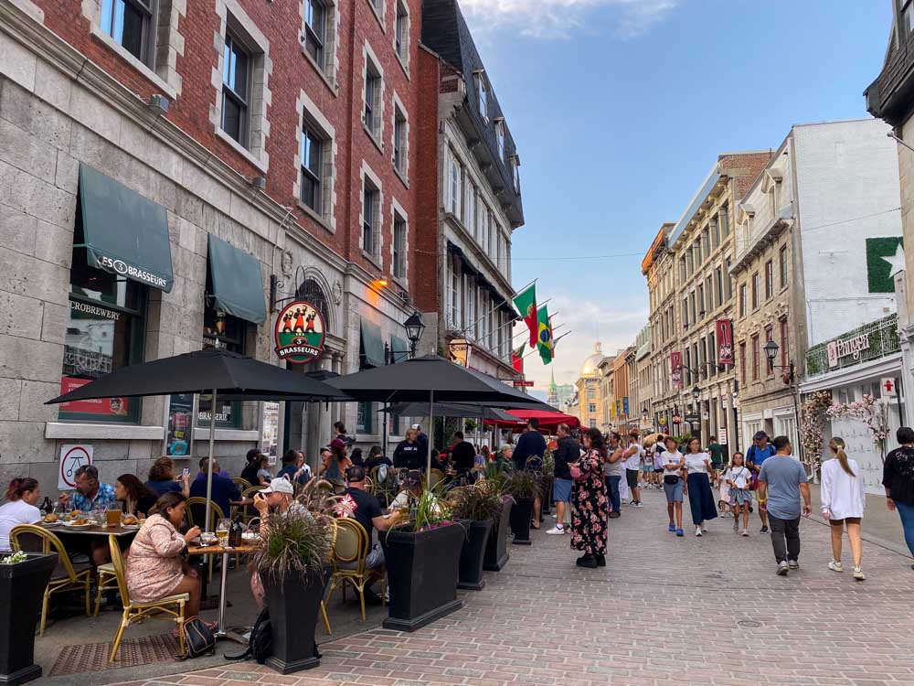 A busy evening on Rue Saint-Paul in Old Montreal with many people walking down the street and eating at sidewalk tables. Bonsecours Market is visible in the background.
