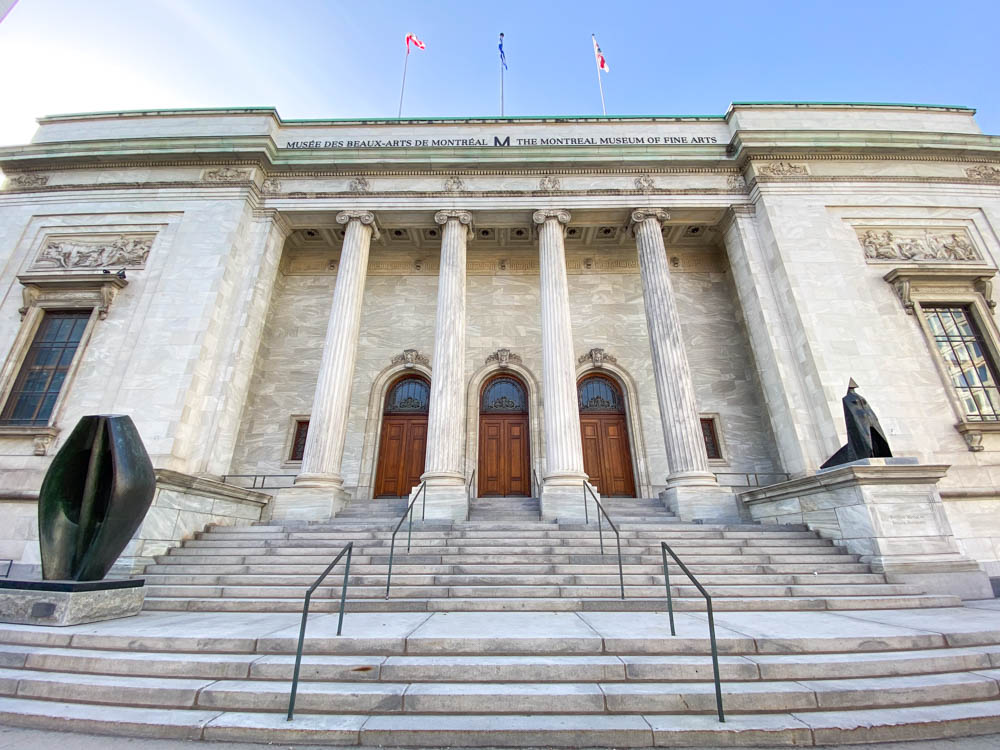 Facade of the Michal and Renata Hornstein pavilion (a large stone building with 4 pillars, 3 flags on the roof, and sculptures on either side of the steps) at the Montreal Museum of Fine Arts.