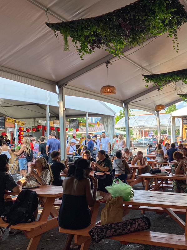Crowds of people eating at picnic tables next to the weekend food stalls at Atwater Market.