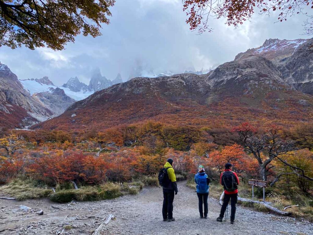 Three hikers on the Laguna de los Tres trail waiting for the clouds around Mount Fitz Roy to clear. The mountain slope in front of them is covered in autumn leaves.