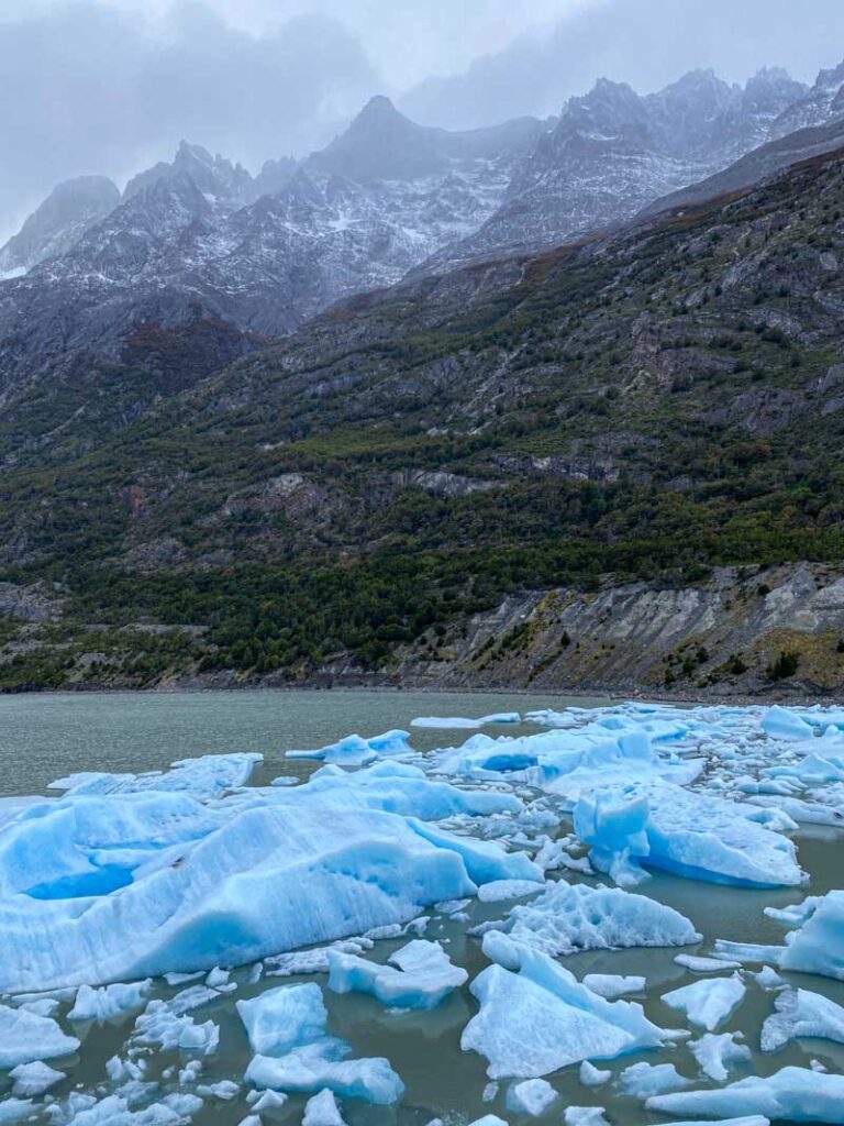 Large blue chunks of ice float in a lake below snowy mountain peaks at Grey Glacier viewpoint about 10 minutes from Refugio Grey.