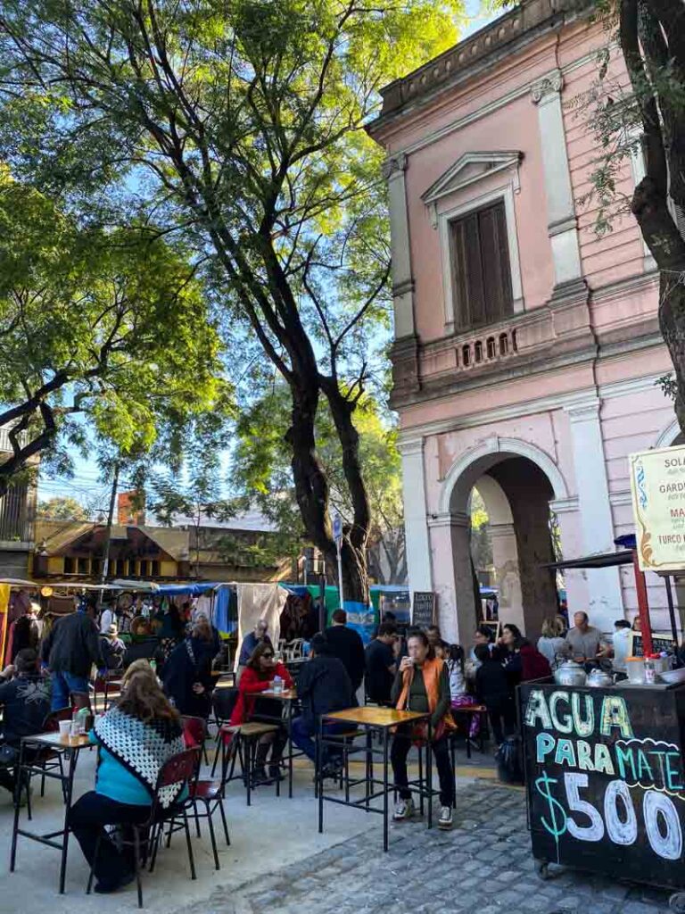 People eat lunch at outdoor tables under tall trees and a pink colonial-style building at the Mataderos Fair in Buenos Aires.