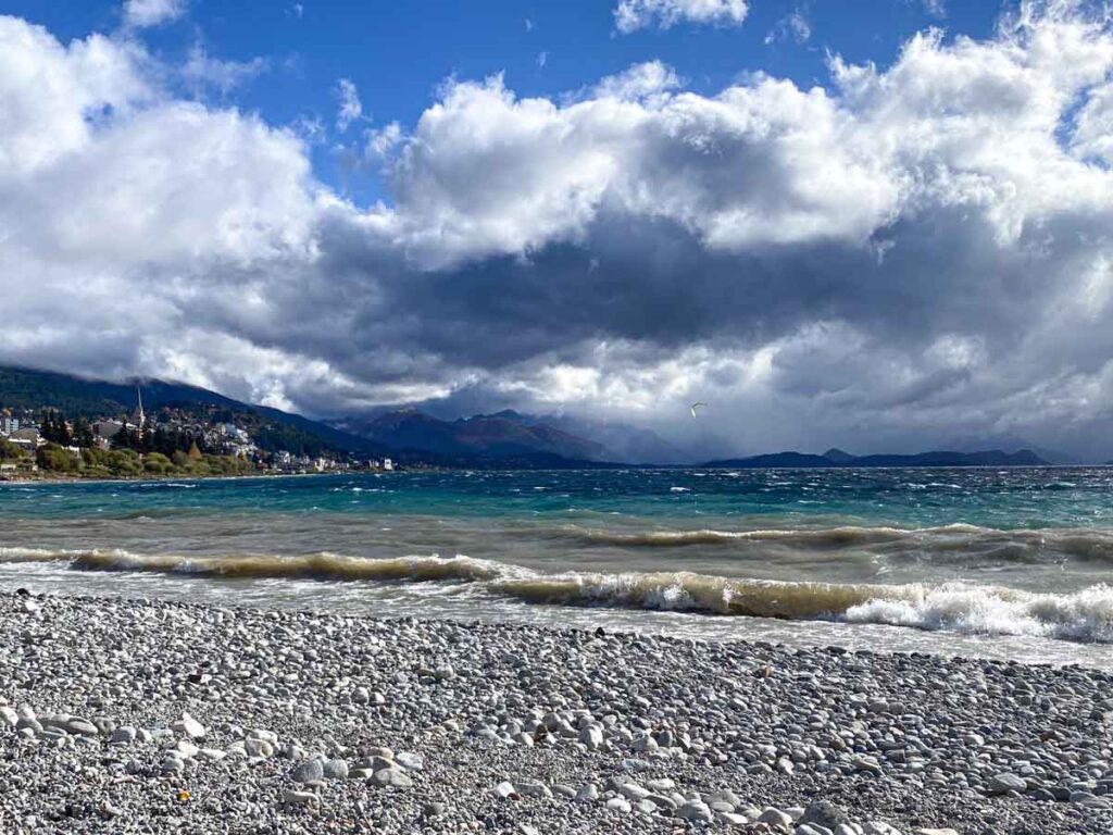 View from the pebble beach at Playa Centenario - Bariloche Cathedral and town center on the left, with mountains and a kitesurfer on Nahuel Huapi Lake to the right.
