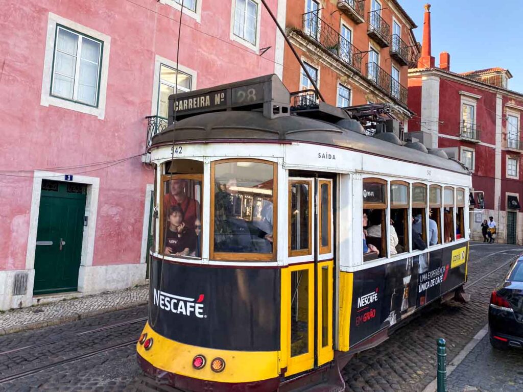 A classic 28E tram packed with tourists traveling along a cobblestone street next to colorful Lisbon buildings.