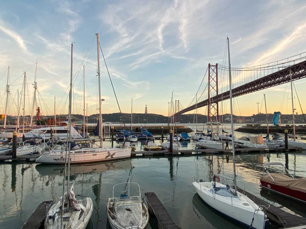 Boats in a harbor on the Tagus River at sunset, just below Ponte 25 de Abril, with the iconic Santuario Cristo Rei visible across the water.