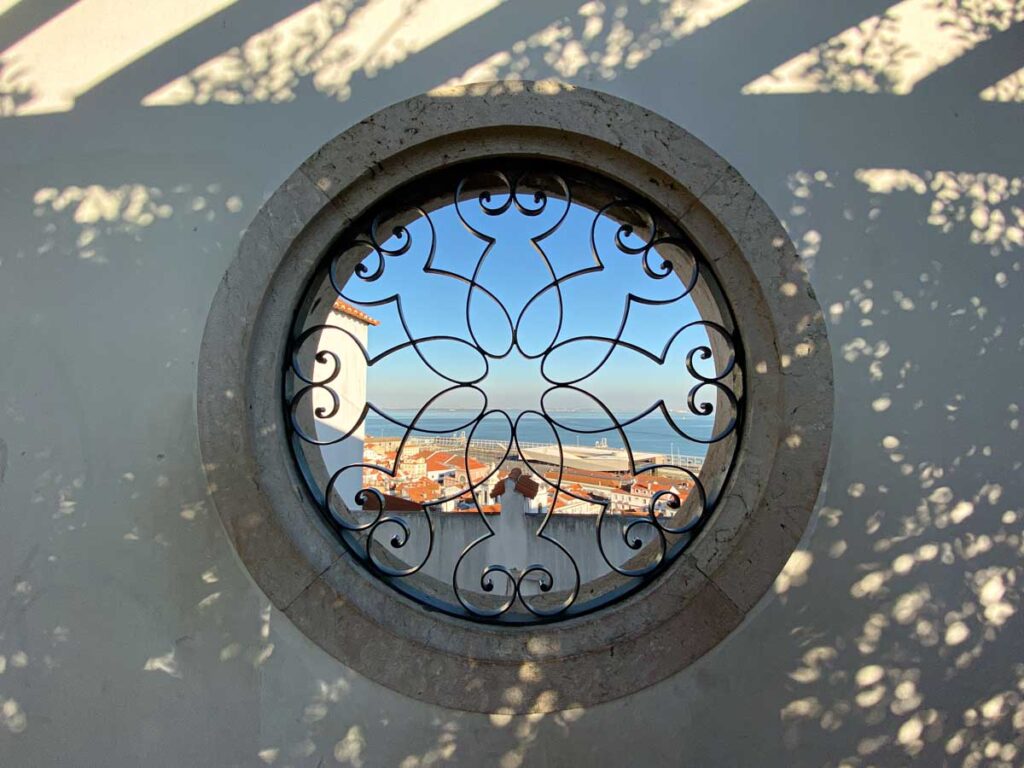 Circular window at Miradouro de Santa Luzia with a view over the Tagus River and the classic orange rooftops of Lisbon.